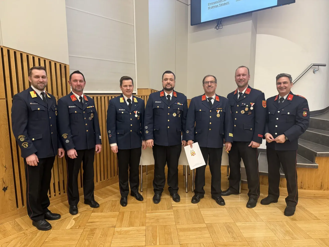 Eine Gruppe uniformierter Männer steht in einem Konferenzraum und lächelt. Sie tragen Medaillen und halten ein Zertifikat. Ein großer Bildschirm ist im Hintergrund.