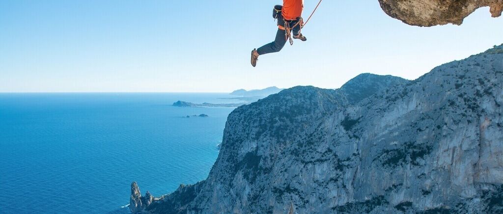Eine Person in einem orangefarbenen Shirt und schwarzen Hosen klettert an einem Seil hängend. Darunter befindet sich der Ozean.
