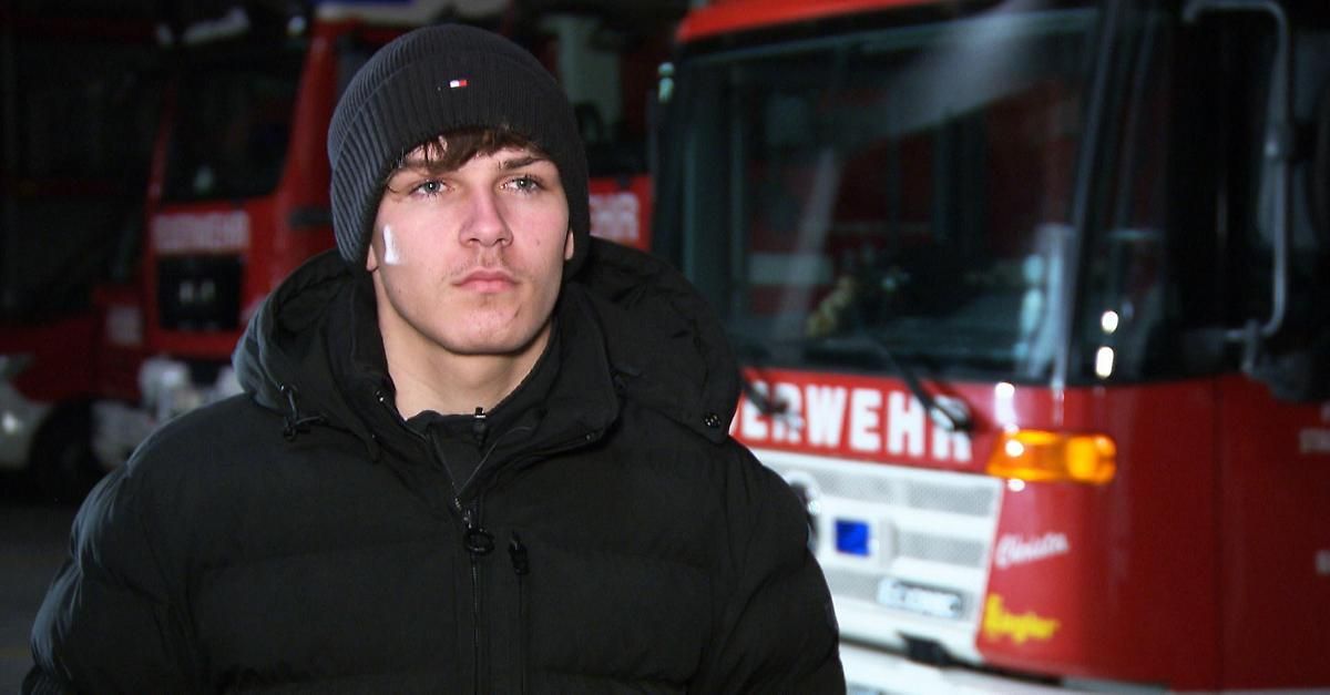 Ein Mann in einer schwarzen Mütze und Jacke steht vor einem roten Feuerwehrauto. Das Auto hat Text und Logos auf der Front.