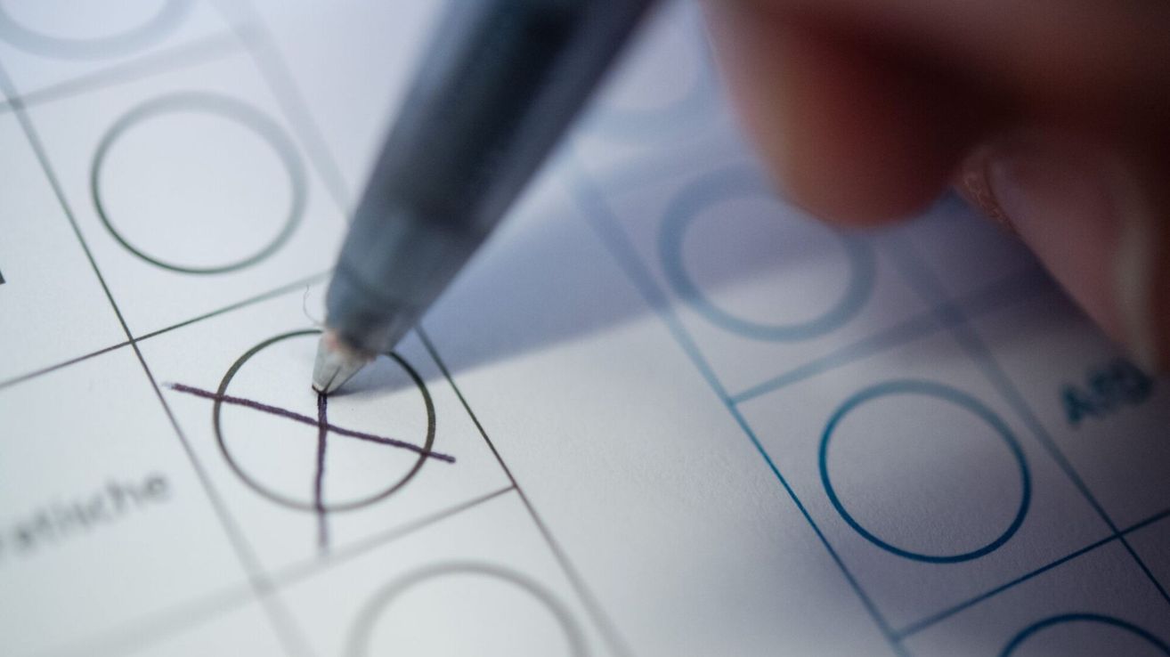 Eine Hand hält einen Stift über ein Papier mit Kontrollkästchen, was darauf hinweist, dass ein Formular ausgefüllt wird. Das Papier hat mehrere Kreise und ein Kreuzmarkierung.