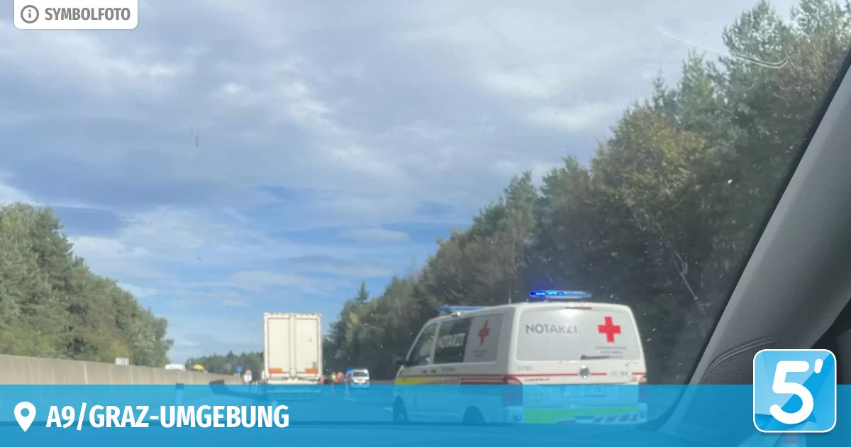 Ein Krankenwagen mit eingeschaltetem Blaulicht steht am Straßenrand, mit einem Lkw dahinter. Bäume und ein blauer Himmel mit Wolken sind im Hintergrund zu sehen.