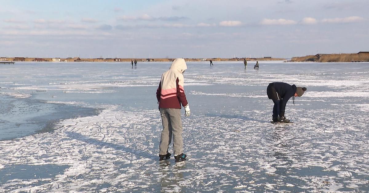Eine Gruppe von Menschen, die auf einem zugefrorenen See Schlittschuh laufen, wobei eine Person in der Nähe der Kante gebeugt ist.