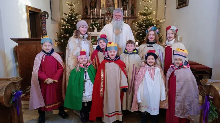 Eine Gruppe von Kindern in traditionellen Kostümen steht mit einem Priester vor einem geschmückten Weihnachtsbaum. Der Priester lächelt und posiert für ein Foto.