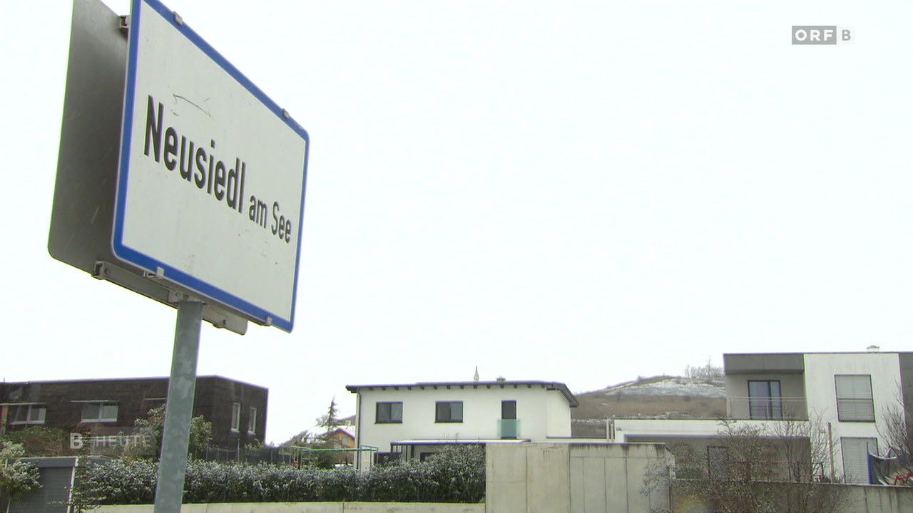 Ein Schild mit der Aufschrift 'Friedl am See' mit einem Haus und Bäumen im Hintergrund. Das Haus hat ein grünes Geländer und mehrere Fenster.