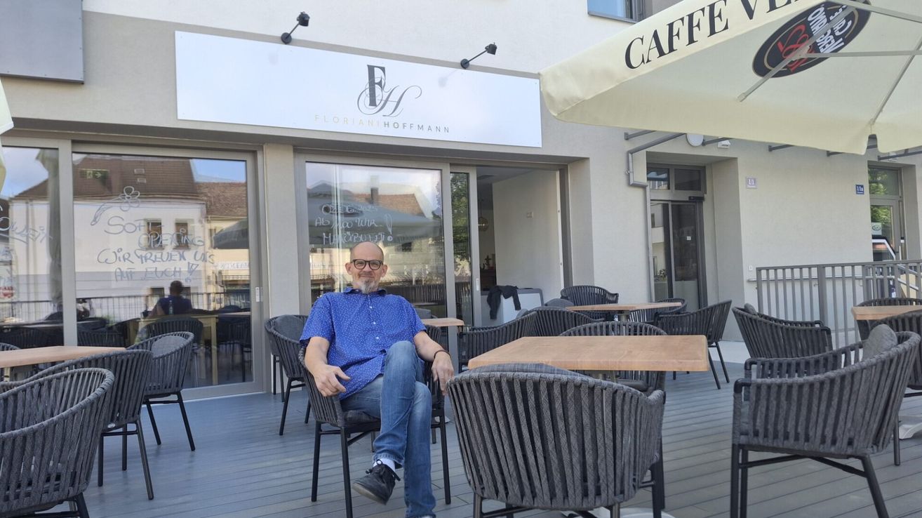 Ein Mann mit Brille sitzt auf einem Stuhl vor einem Restaurant. Das Restaurant hat eine Glastür und Fenster.