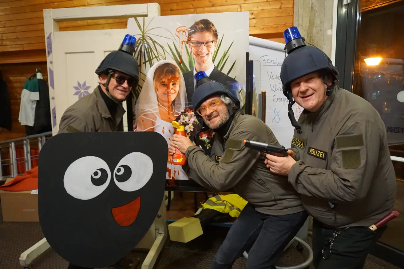 Drei Personen in Polizeiuniformen halten Pistolen, eine mit Brille und Helm, stehen vor einem Bild von Braut und Bräutigam mit roter Farbe.