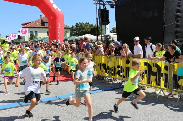 Kinder nehmen an einem Marathon teil, begleitet von einer jubelnden Menge. Sie laufen auf einer Straße mit einem gelben Banner, während Zuschauer hinter einem Zaun zusehen.