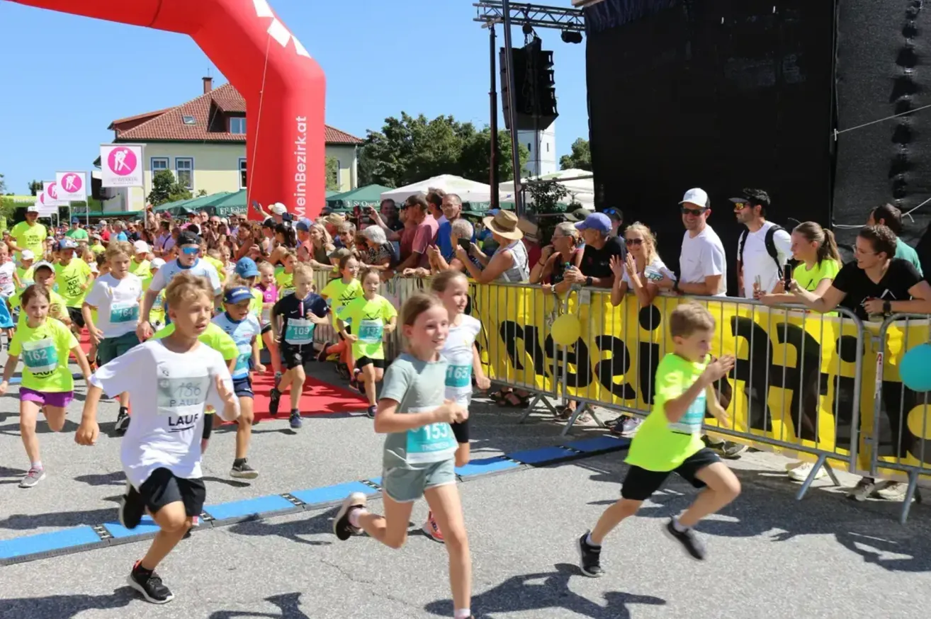 Kinder nehmen an einem Marathon teil, begleitet von einer jubelnden Menge. Sie laufen auf einer Straße mit einem gelben Banner, während Zuschauer hinter einem Zaun zusehen.