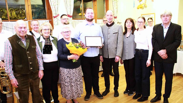 Eine Gruppe von Menschen steht auf einem Holzboden und posiert für ein Foto. Ein Mann hält eine gerahmte Urkunde und eine Frau einen Blumenstrauß. Hinter ihnen stehen zwei Frauen nebeneinander. Dahinter stehen zwei Männer, und ein Mann steht neben einer Glastür.