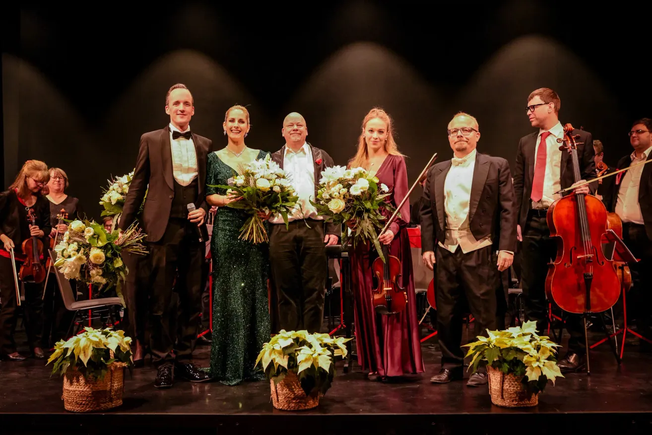 Sechs Personen in formeller Kleidung stehen auf der Bühne und halten Blumen. Zwei Frauen halten Blumensträuße, und ein Mann mit einer Geige steht zwischen ihnen.