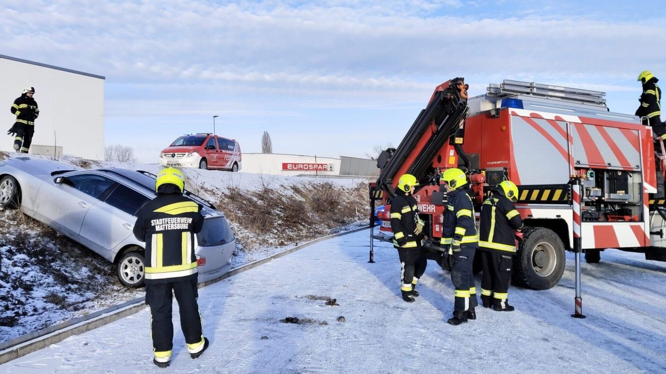 Feuerwehrleute in Uniform stehen auf verschneiten Boden in der Nähe eines roten Feuerwehrwagens. Ein roter Van ist auf der Straße geparkt. Hinter den Feuerwehrleuten befindet sich ein Gebäude mit einem Schild, auf dem EuroSpar steht.