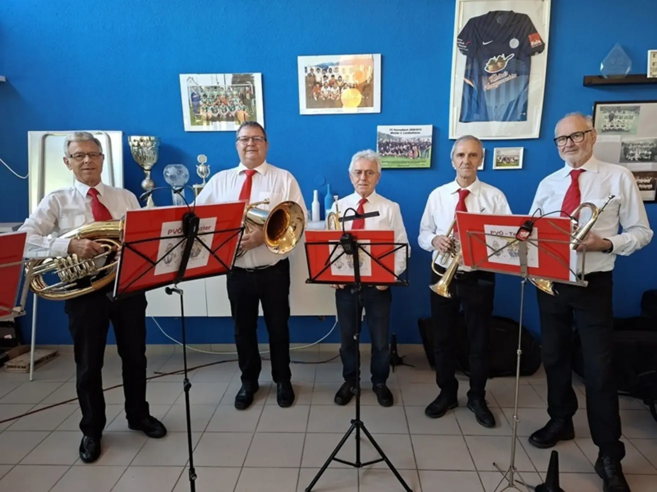 Fünf Männer in roten Krawatten und Brillen stehen vor einer blauen Wand mit gerahmten Bildern und halten Blechblasinstrumente. Sie sind wahrscheinlich Musiker in einer Probe.