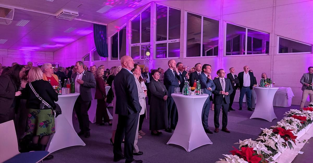 Eine Indoor-Veranstaltung mit formeller Kleidung, bei der die Teilnehmer sich unterhalten und Getränke genießen. Der Raum ist mit lila Lichtern beleuchtet und es gibt große Fenster.