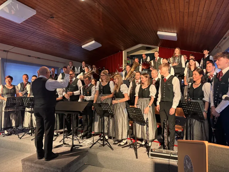 Eine Gruppe von Musikern, darunter sowohl Männer als auch Frauen, steht mit ihren Instrumenten auf der Bühne, wahrscheinlich in Vorbereitung auf eine Aufführung. Der Dirigent zeigt auf die Musiker. Dahinter stehen weitere Musiker, und im Hintergrund befinden sich Holzbalken.