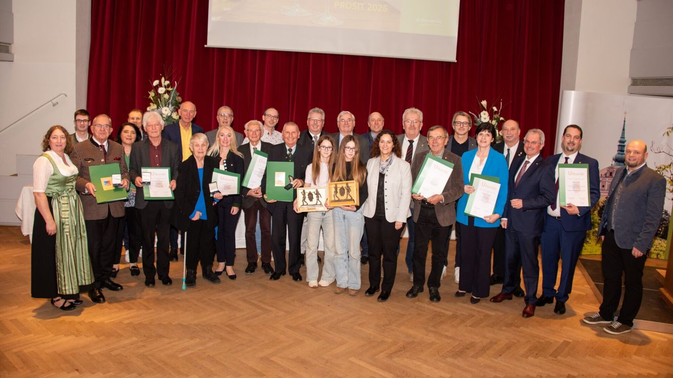 Eine Gruppe von Menschen posiert für ein Foto bei einer Preisverleihung. Sie stehen vor einem roten Vorhang und halten Urkunden. Der Mann und die Frau in der Mitte halten gerahmte Auszeichnungen.