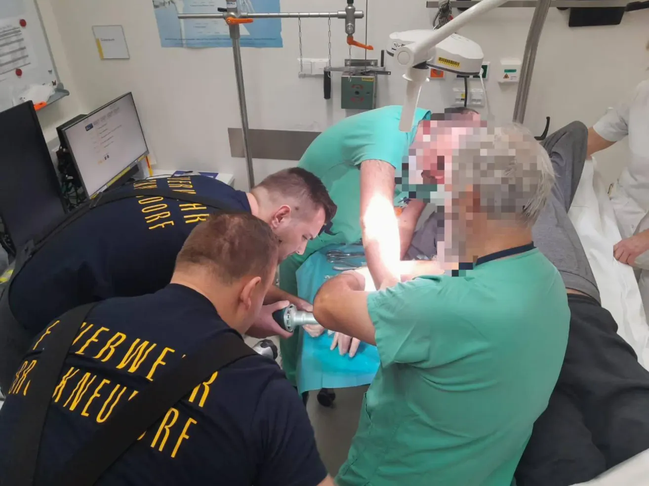 Eine Gruppe medizinischer Fachkräfte führt einen Eingriff in einem Krankenhauszimmer durch, wobei ein Patient auf dem Tisch liegt und verschiedene medizinische Instrumente sichtbar sind.