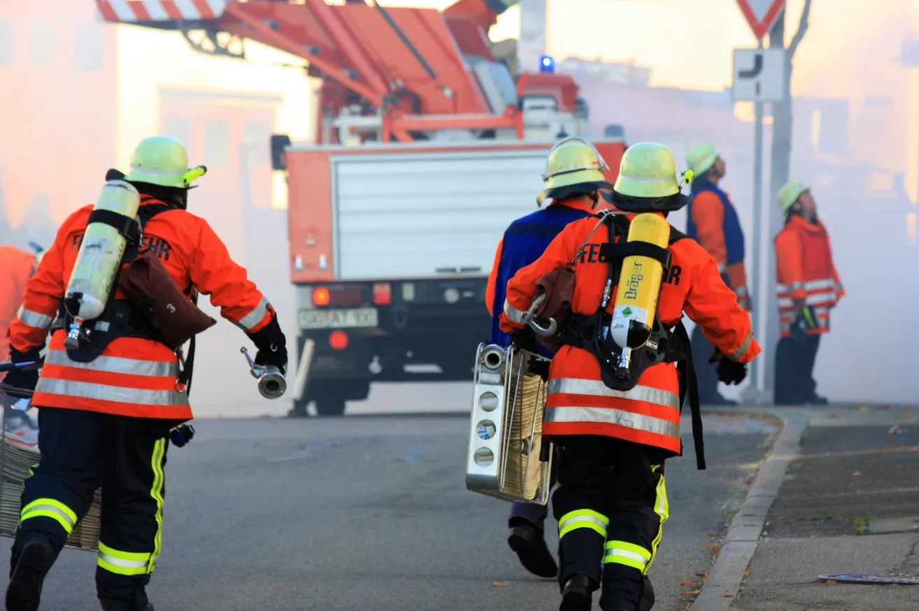 Eine Gruppe von Feuerwehrleuten mit Helmen und Schutzanzügen geht mit Ausrüstung auf einer Straße. Dahinter ist ein Feuerwehrwagen geparkt.