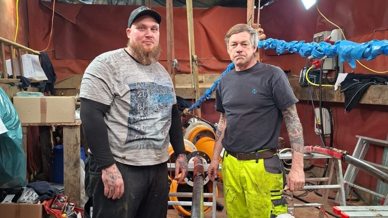 Zwei Männer in einer Fabrik, einer trägt eine Kappe und ein graues T-Shirt, der andere hat Tattoos und ein schwarzes T-Shirt. Sie stehen neben einer großen Rohrleitung.