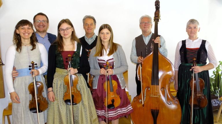 Eine Gruppe von Musikern posiert für ein Foto. Zwei Frauen halten Geigen, während ein Mann rechts ein Cello hält. Die Männer tragen Westen und die Frauen traditionelle Kleider.