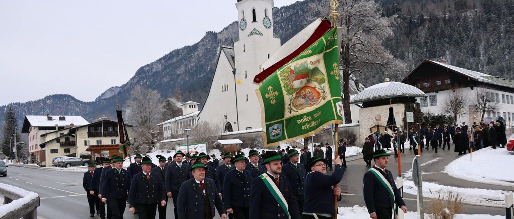Eine Gruppe von Menschen in Uniformen und Hüten marschiert mit einer Fahne und einem Pfahl vor einer Kirche auf einer verschneiten Straße.