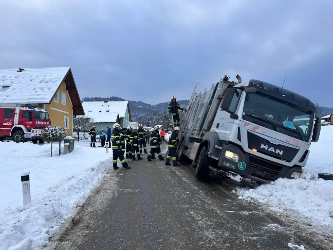 Ein umgestürzter Lastwagen auf einer verschneiten Straße, mit Feuerwehrleuten in der Nähe. Die Szene spielt sich in einer verschneiten Landschaft mit Häusern im Hintergrund ab.