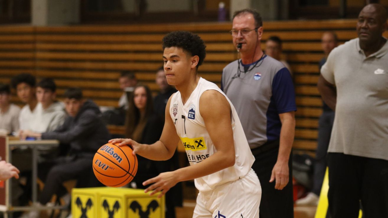 Ein Basketballspieler in einem weißen Trikot mit der Nummer 7 dribbelt den Ball während eines Spiels, während ein Schiedsrichter hinter ihm steht.