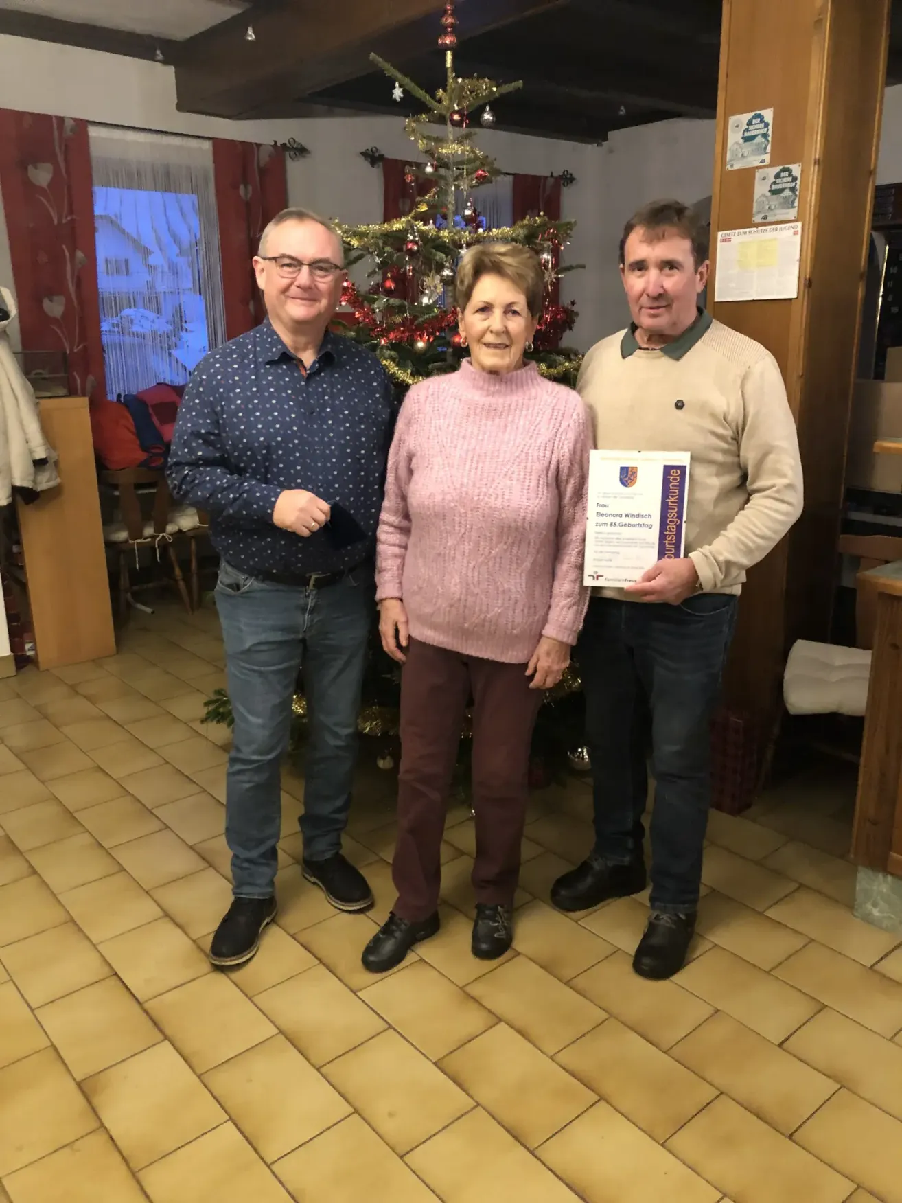 Drei Menschen stehen lächelnd nebeneinander vor einem Weihnachtsbaum. Die Frau in der Mitte hält ein Zertifikat.