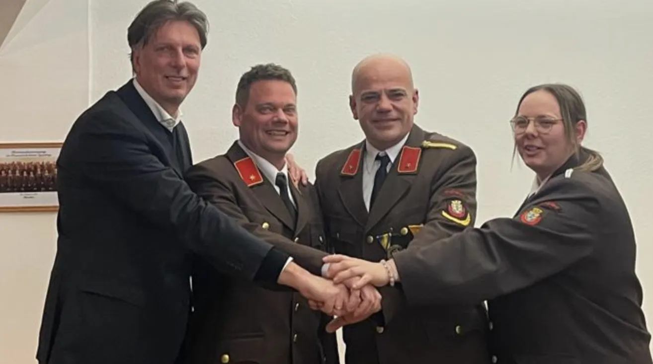 Vier Männer in formellen Uniformen, zwei in Militäruniformen, einer in einem schwarzen Anzug und ein weiterer in einem weißen Hemd, schütteln Hände. Sie scheinen sich auf einer Veranstaltung oder einem Treffen zu befinden.
