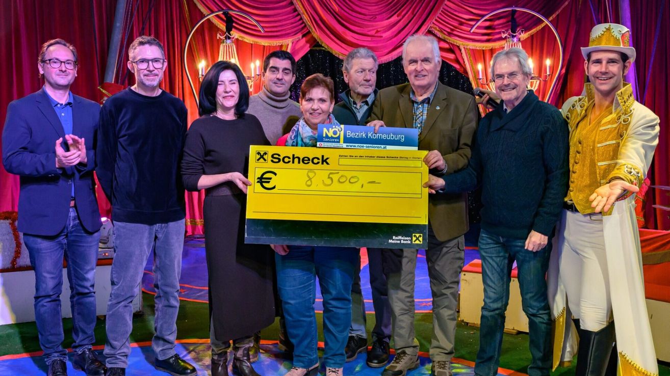 Eine Gruppe von Menschen steht auf einer Bühne und hält einen großen gelben Scheck über 8.500 Euro, gesponsert von Raiffeisen Meine Bank, vor einem Hintergrund aus roten Vorhängen und einem Kronleuchter.