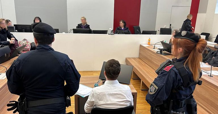 Ein Mann sitzt in einem Gerichtssaal und sieht den Richtern gegenüber. Zwei Polizeibeamte stehen in der Nähe, einer mit einem Mikrofon. Die Richter sitzen, und eine rote Wand ist hinter ihnen.