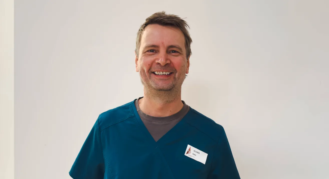 Ein Mann in blauer Arbeitskleidung lächelt in die Kamera. Er trägt einen Namensschild mit dem Namen Oliver.