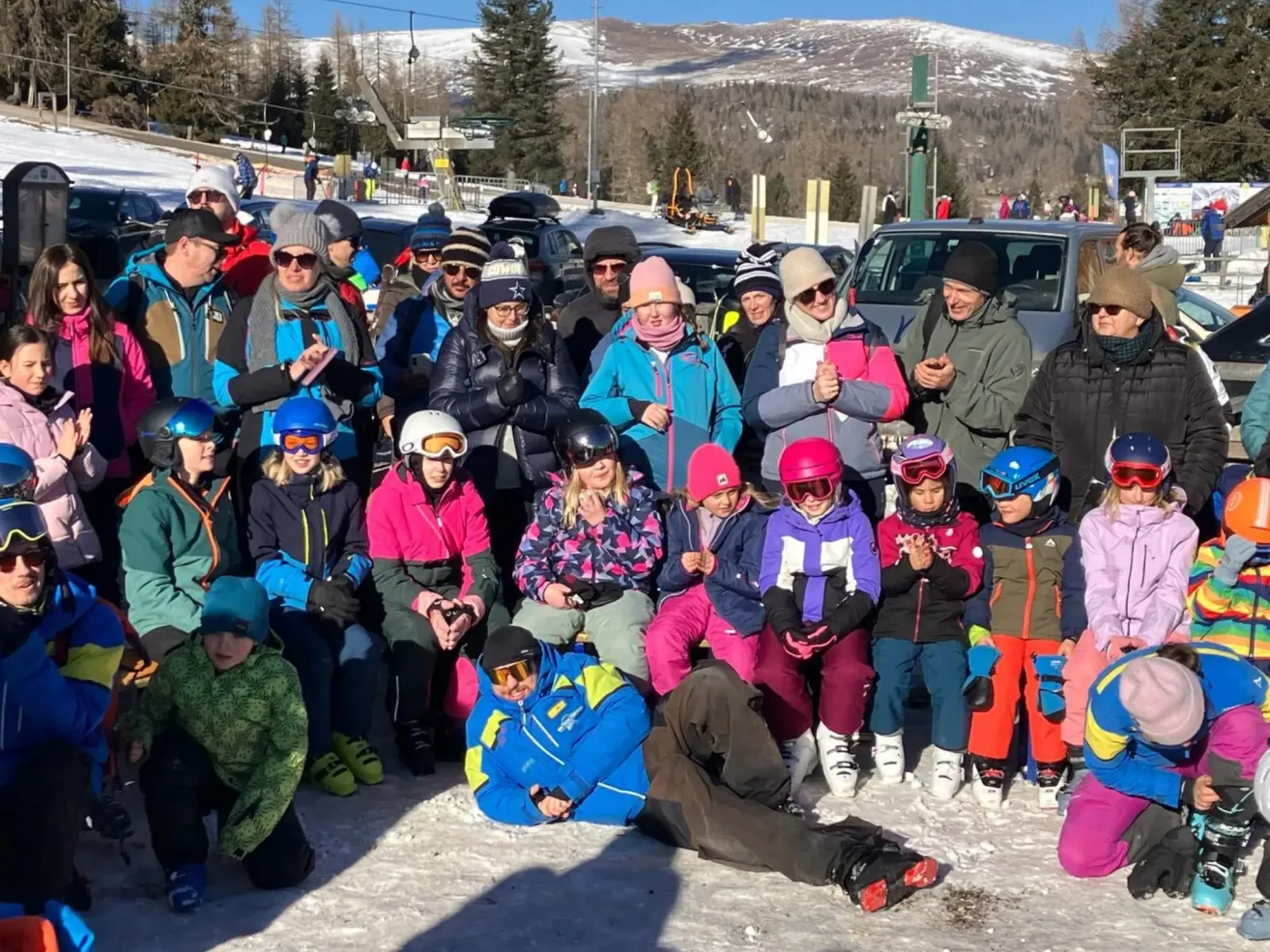 Eine Gruppe von Menschen, einige in Winterkleidung, versammelt sich in einem verschneiten Außenbereich. Einige sitzen, andere stehen. Der schneebedeckte Berg und die Bäume sind im Hintergrund.
