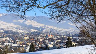 Eine verschneite Landschaft mit einer verschneiten Stadt, die verschiedene Gebäude, eine Kirche und Berge im Hintergrund zeigt.