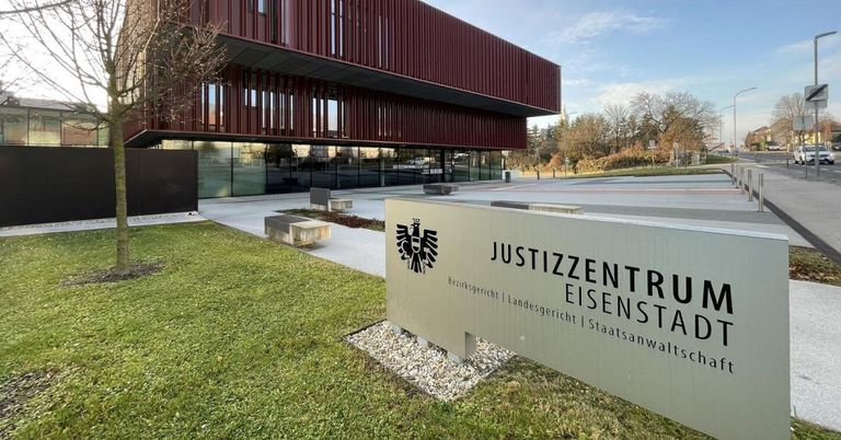 Ein modernes Gebäude mit roter Metallfassade und Glaswänden ist zu sehen. Ein Schild davor steht 'JUSTIZZENTR EISEN'. Die Szene ist im Freien mit Bäumen und klarem Himmel.