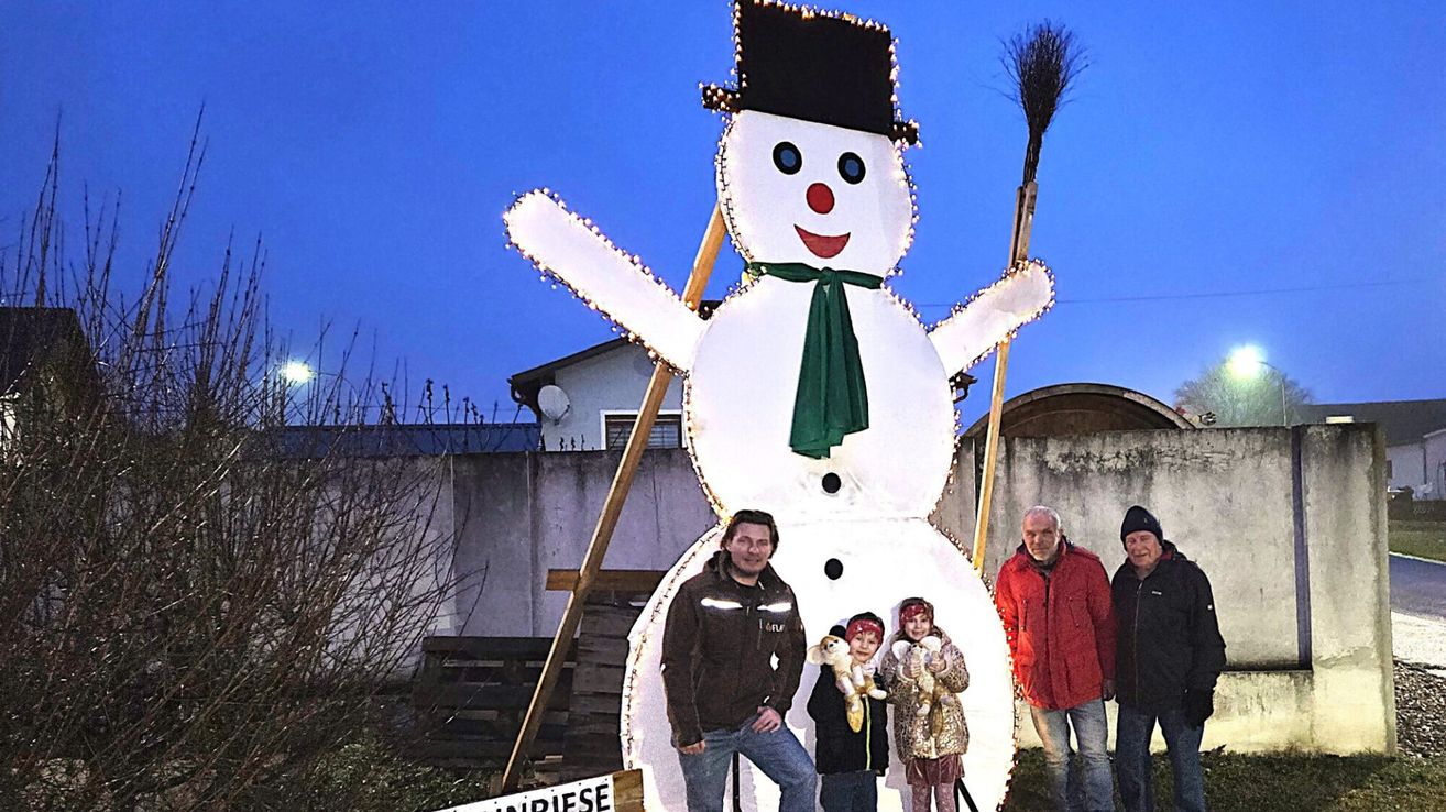 Eine vierköpfige Familie, darunter zwei Kinder, steht vor einem großen, beleuchteten Schneemann. Der Schneemann ist mit Lichtern dekoriert und hat eine Besen in der Nähe. Sie sind draußen, mit einer Wand und einigen Bäumen im Hintergrund.