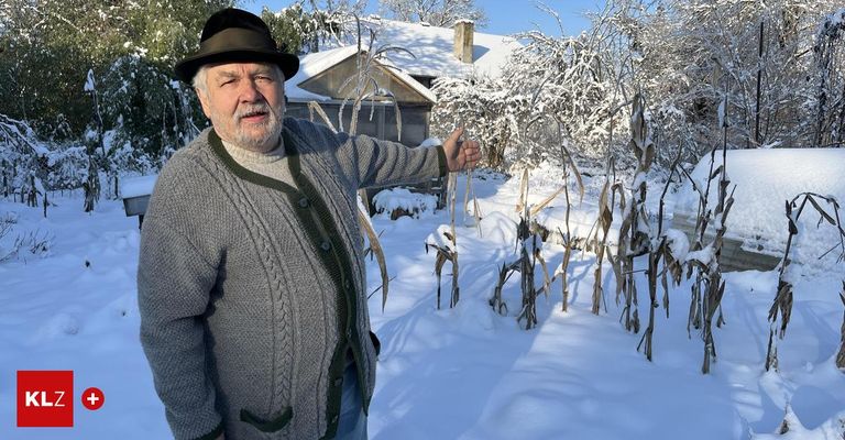 Ein älterer Mann in einem Hut und einer grauen Strickjacke steht in einem verschneiten Garten und zeigt auf einige hohe, vertrocknete Pflanzen. Hinter ihm ist ein Haus mit Schnee auf dem Dach zu sehen.