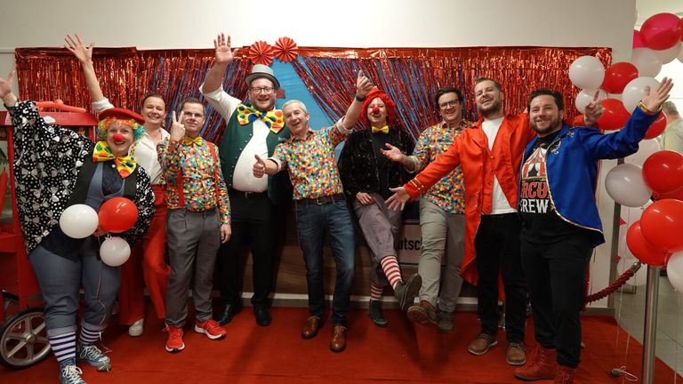 Eine Gruppe von Menschen in bunten Clown-Kostümen posiert für ein Foto vor einem roten Vorhang mit rotem Teppich.