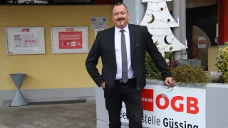 Ein Mann in Anzug steht vor einem Weihnachtsbaum und einem Schild, das 'OGG' steht. Er lächelt und hat die Hände in den Taschen.