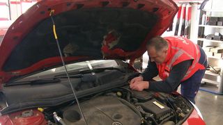 Ein Mann in einer Warnweste arbeitet mit offenem Motorhauben an einem Automotor. Er hält ein Werkzeug in der Nähe der Batterie.