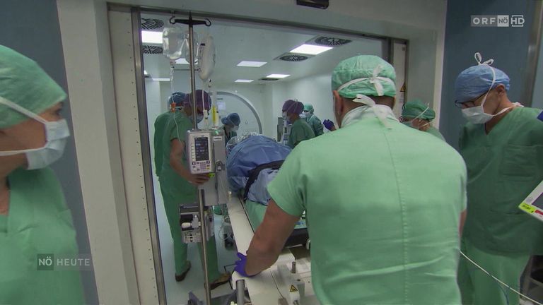 Medizinisches Personal in grünen Scrubs bereitet einen Patienten in einem Operationssaal mit offener Tür auf eine Operation vor. Geräte und Monitore sind sichtbar.