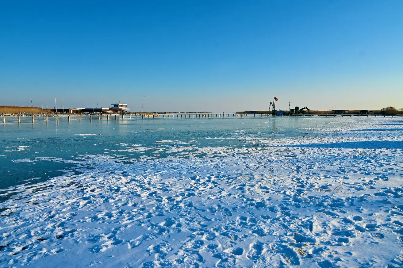 Eine verschneite Landschaft mit einem Steg in der Ferne. Der Himmel ist klar, und es gibt einige Maschinenfahrzeuge auf dem Steg.