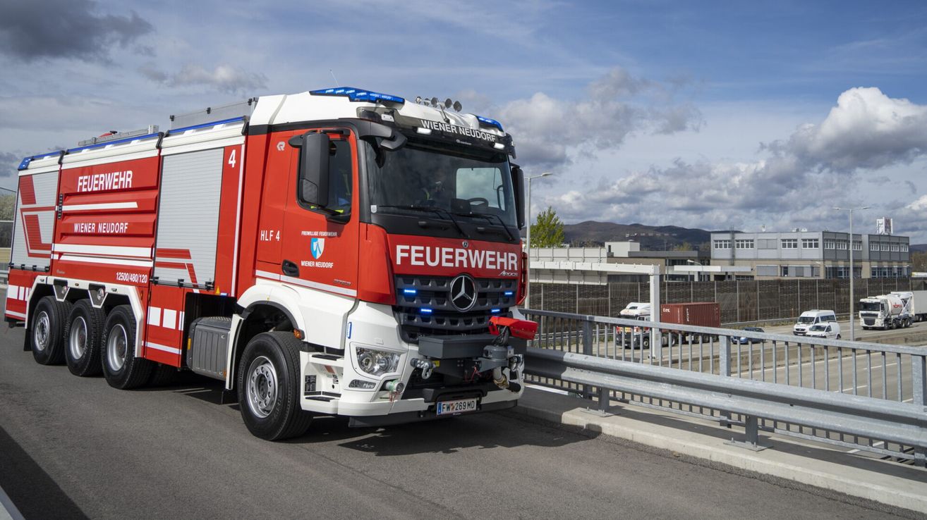 Ein roter und weißer Mercedes-Benz Feuerwehrwagen, beschriftet mit 'Wiener Neudorf', fährt auf einer Straße mit einer Metallbarriere und entfernten Gebäuden.
