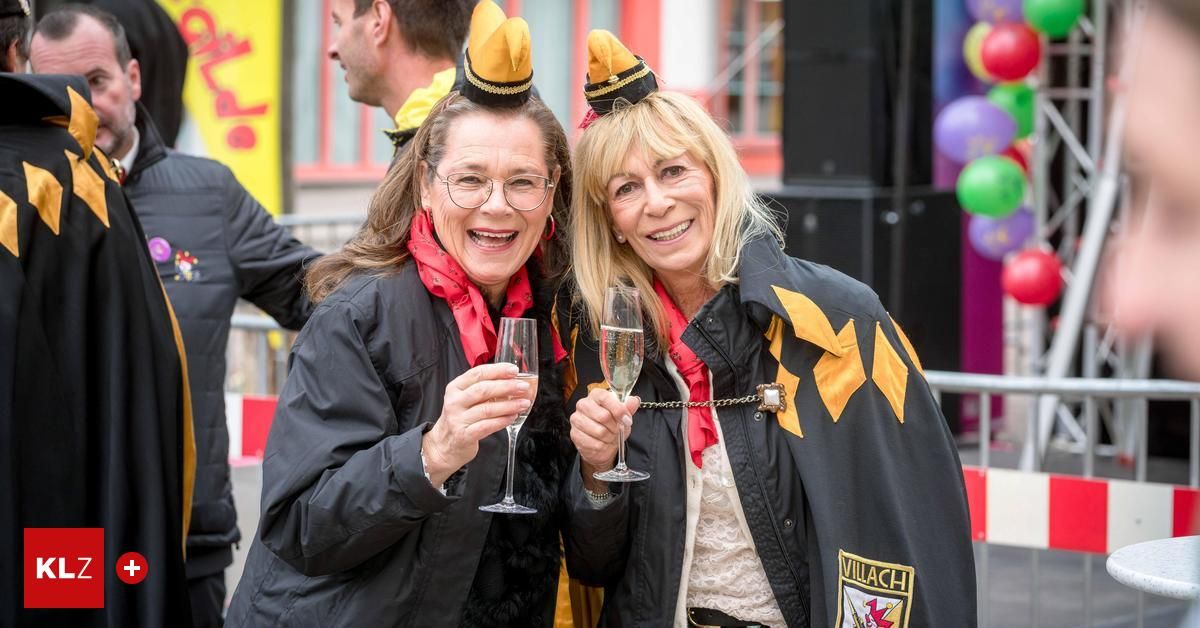 Zwei Frauen in leuchtenden Kostümen, eine mit einem roten Schal, halten Champagnergläser und lächeln für ein Foto bei einer festlichen Veranstaltung.