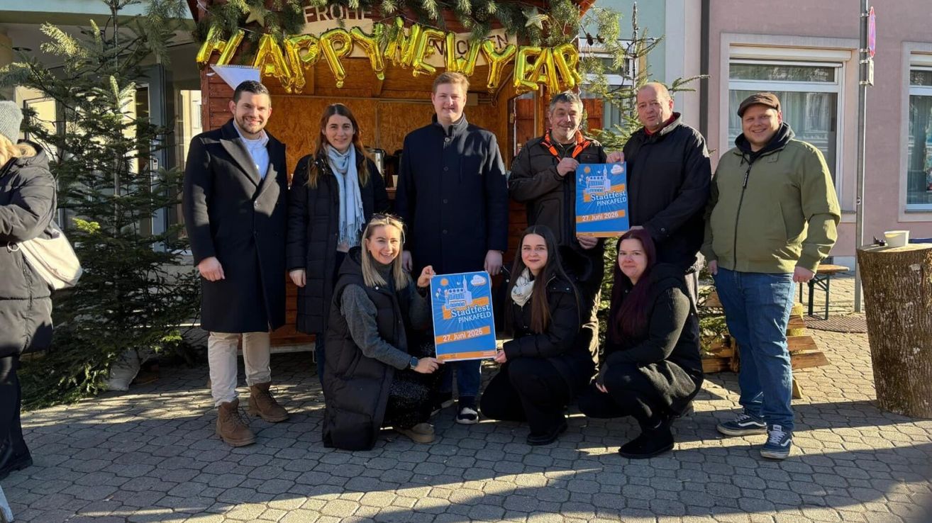 Eine Gruppe von Menschen in Winterkleidung posiert für ein Foto vor einem Stand mit einem Happy New Year-Banner und einem Stadtfest Pinkafeld-Poster.