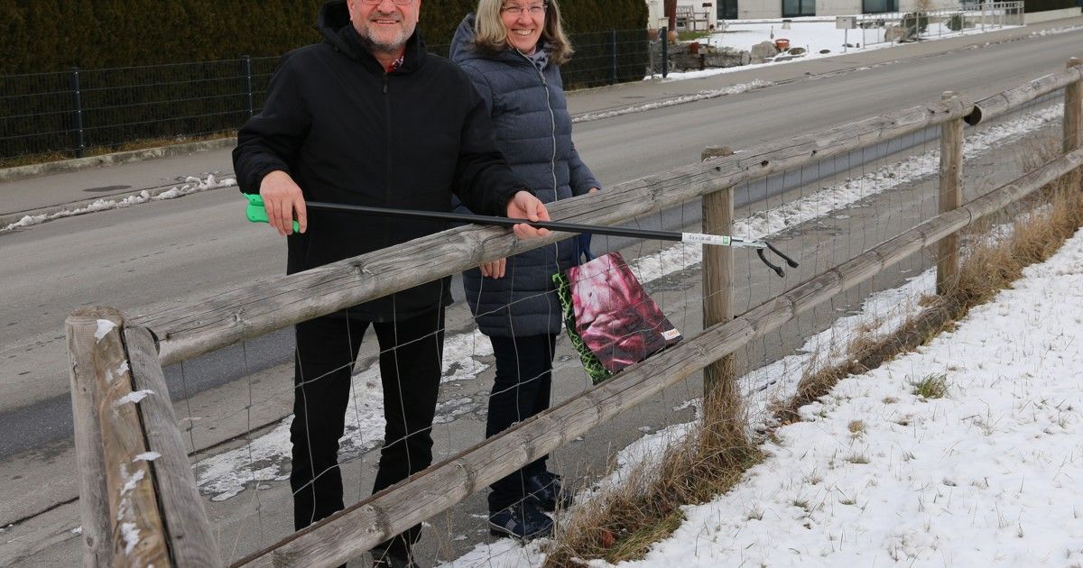 Zwei Personen stehen neben einem Holzzaun und lächeln. Der Mann hält einen langen schwarzen Stock, während die Frau eine Tasche in der Hand hat. Schnee liegt auf dem Boden.
