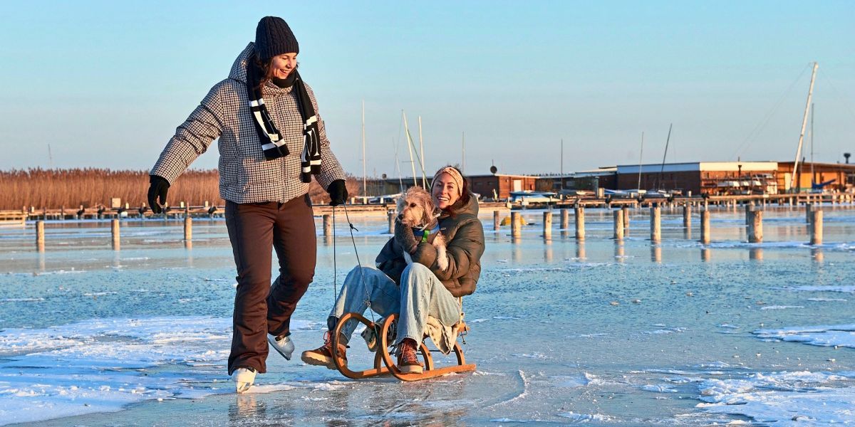 Eine Frau zieht einen Schlitten auf Eis mit einer lächelnden Frau und einem Hund im Inneren, beide in Winterkleidung.