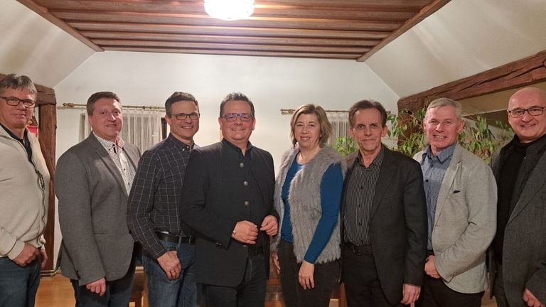 Fünf Personen stehen lächelnd in einem Raum für ein Foto. Sie sind in formeller Kleidung gekleidet, und über ihnen befindet sich eine Holzdecke.