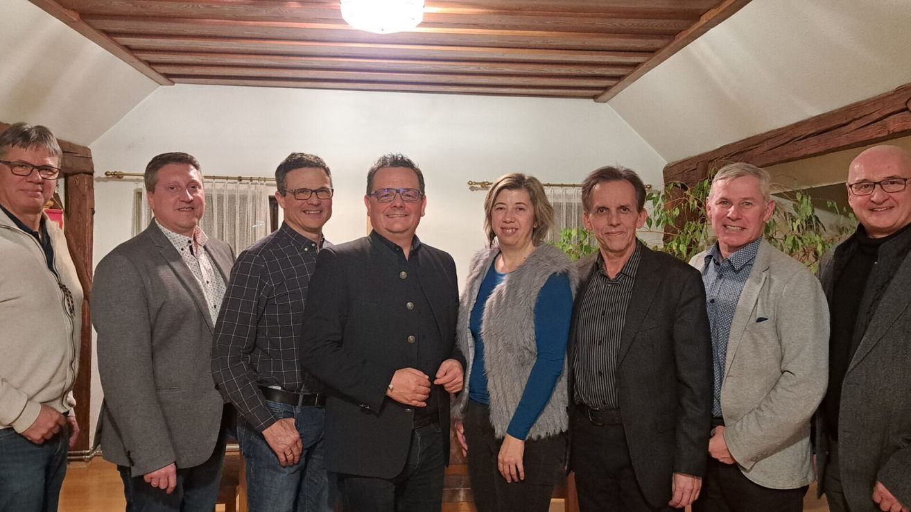 Fünf Personen stehen lächelnd in einem Raum für ein Foto. Sie sind in formeller Kleidung gekleidet, und über ihnen befindet sich eine Holzdecke.