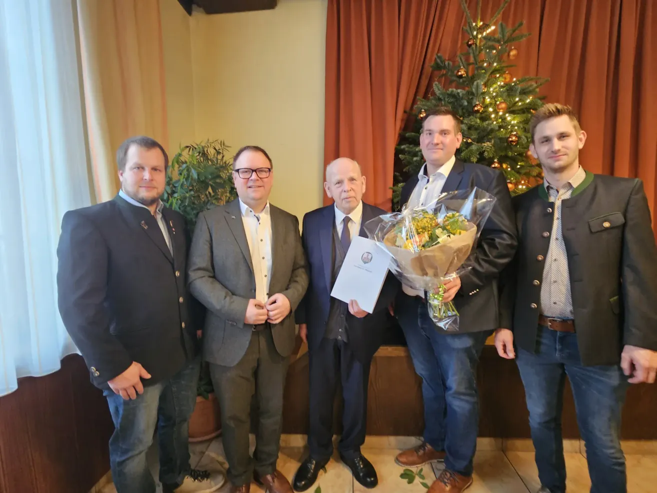 Fünf Männer in Anzügen stehen eng beieinander. Einer hält einen Blumenstrauß und ein Zertifikat. Dahinter steht ein Weihnachtsbaum und ein Fenster mit Vorhängen.