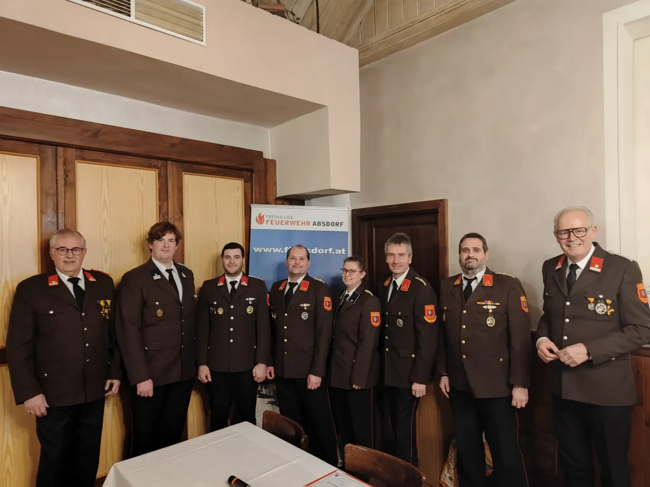 Eine Gruppe von Feuerwehrleuten in Uniform steht vor einem Tisch. Hinter ihnen ist ein Banner mit dem Text 'Feuerwehr Absdof'. Im Hintergrund sind zwei Türen und ein Fenster zu sehen.